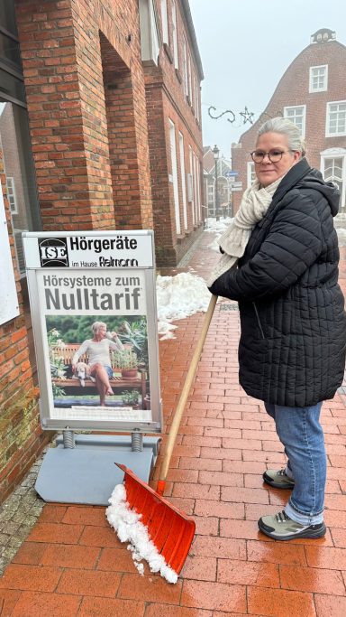 Frau Streich vor der ISE Filiale in Wittmund Frau mit Schneeschaufel vor einem Informationsschild in der Stadt.