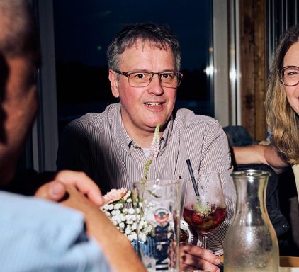 Gesellige Runde am Tisch mit Getränken, ein Mann und eine Frau lächeln.