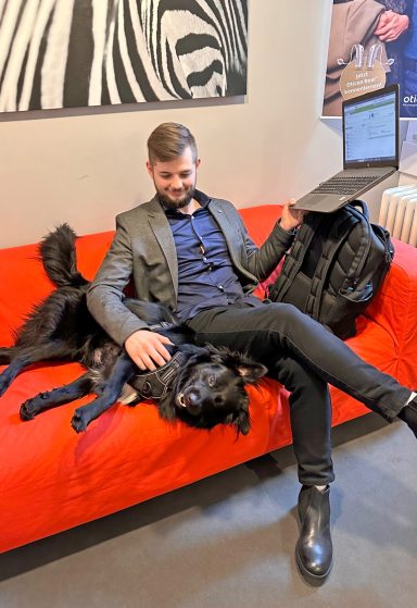 Mann in Anzug sitzt mit Laptop auf rotem Sofa und streichelt einen schwarzen Hund.
