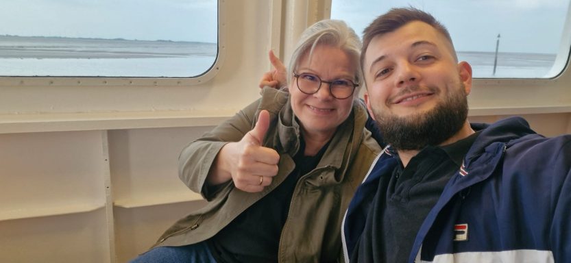 Auf zum Hausbesuch auf die Insel Frau und Mann sitzen an einem Fenster, lächelnd und Daumen hoch.