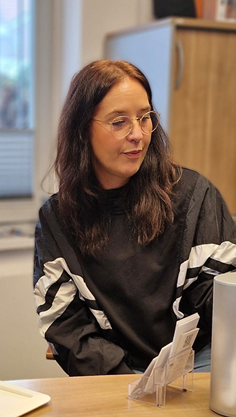 Janna Ise Eine Frau mit langen, braunen Haaren in einem schwarzen Oberteil sitzt an einem Tisch.