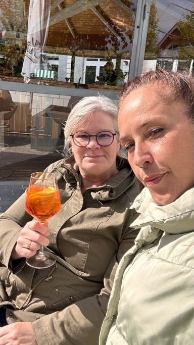 Zwei Frauen, eine hält ein Getränk, sitzen entspannt im Freien.