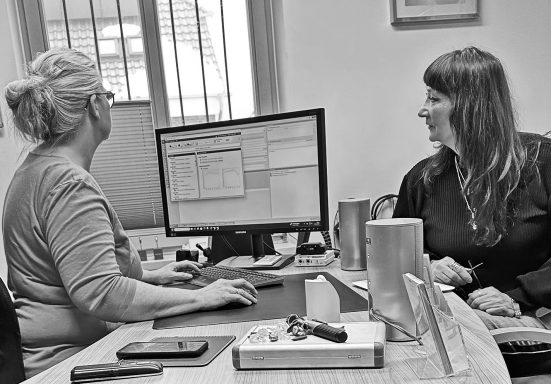 Zwei Frauen sitzen am Tisch, eine arbeitet am Computer, die andere schaut zu.