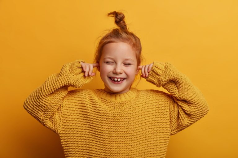 Kinder-Audiologie Lachendes Mädchen in einem gelben Pullover mit einer orangefarbenen Hintergrundfarbe.
