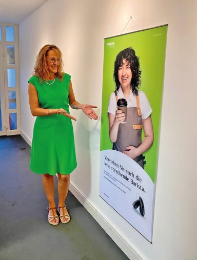Eine Frau in grünem Kleid steht neben einem Werbebanner mit einem lächelnden jungen Mann.